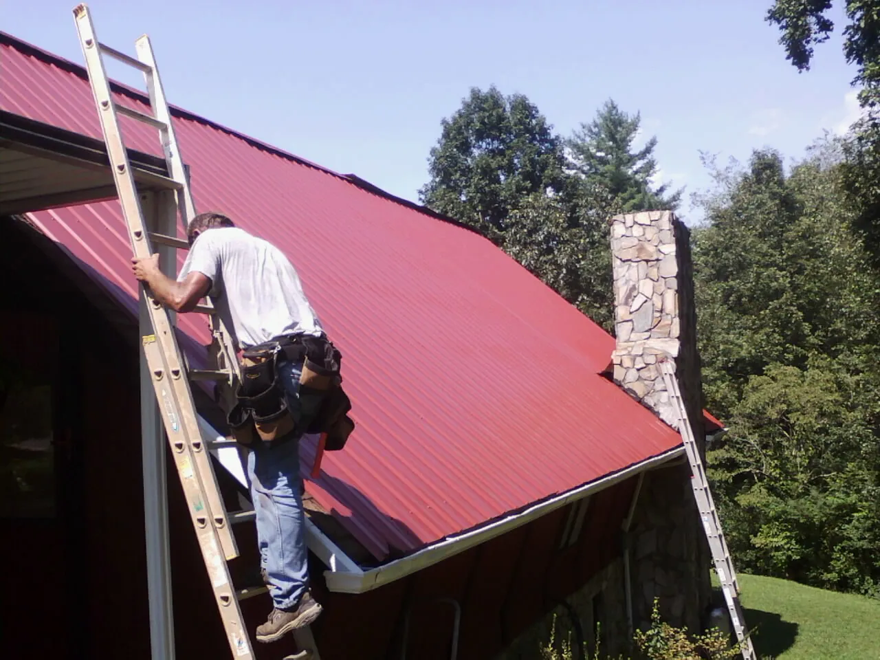 Roofer on a ladder beside a red metal roof and stone chimney — Soffit & Fascia Repair by Roofing Brothers in Lloyd